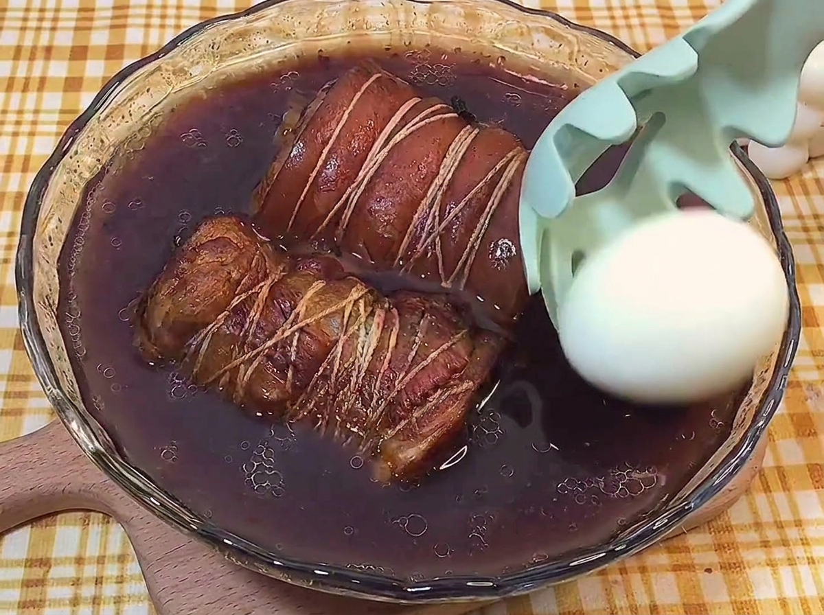 Un huevo cocido pelado siendo sumergido con una espumadera en el líquido oscuro junto a los rollos de chashu.