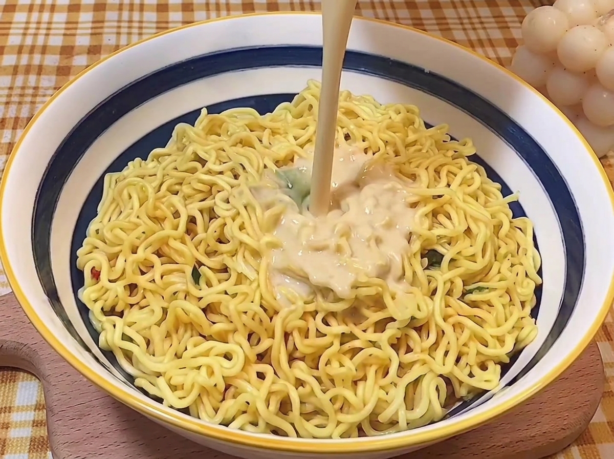 Un chorro de caldo tonkotsu rico y cremoso vertiéndose sobre los fideos cocidos en un tazón.