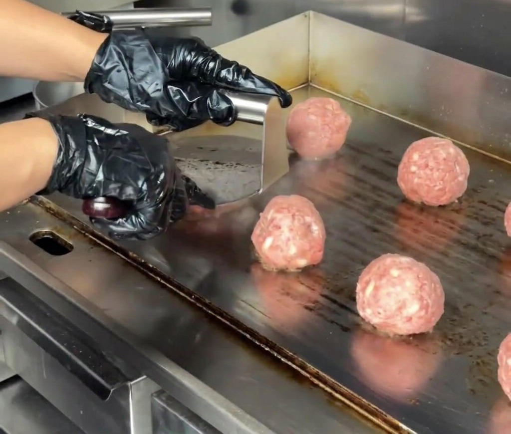 Primer plano de una prensa de acero inoxidable para hamburguesas aplastando una bola de carne hasta formar un medallón grueso en una plancha comercial.