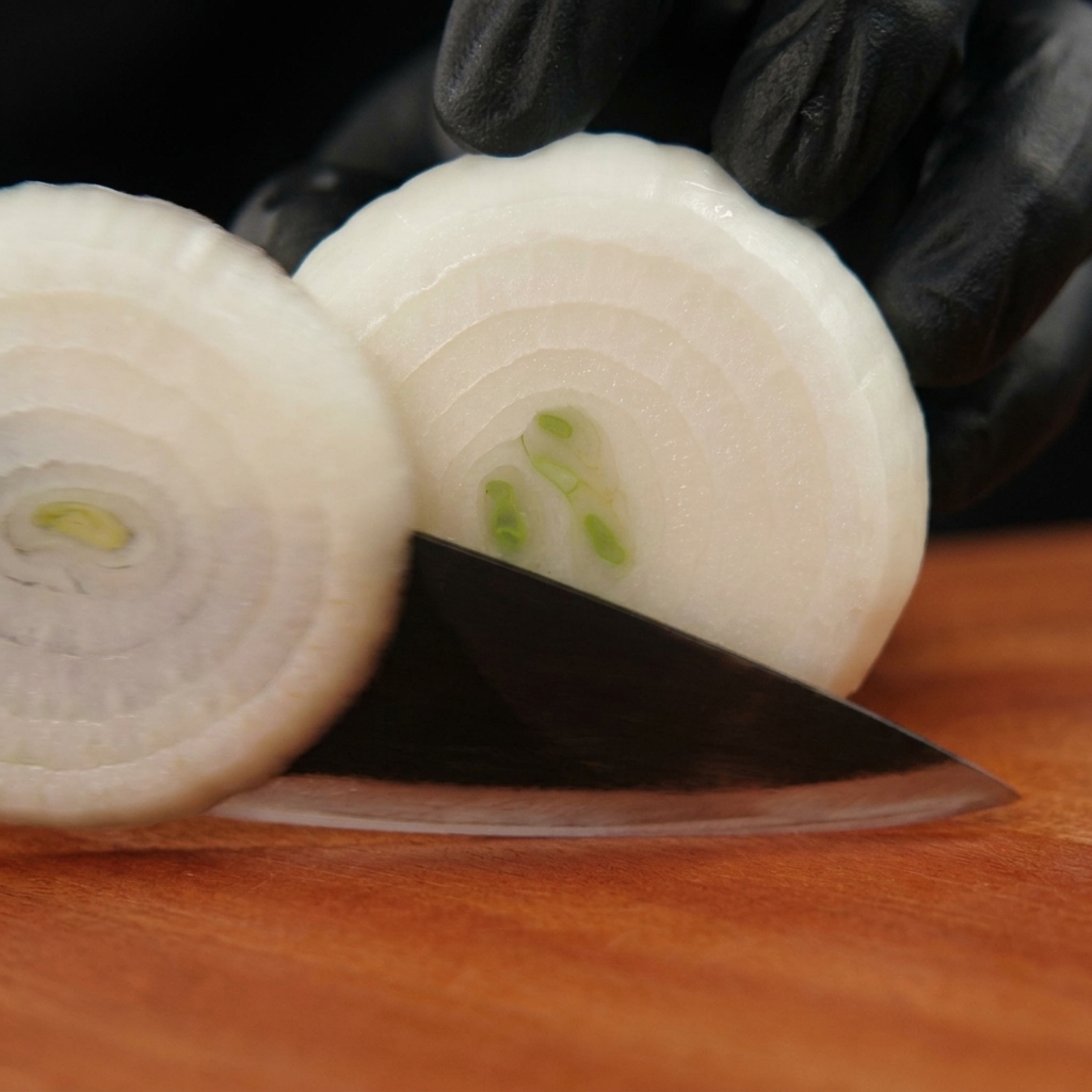 Manos con guantes negros utilizando un cuchillo para cortar una cebolla blanca pelada en aros sobre una tabla de madera.