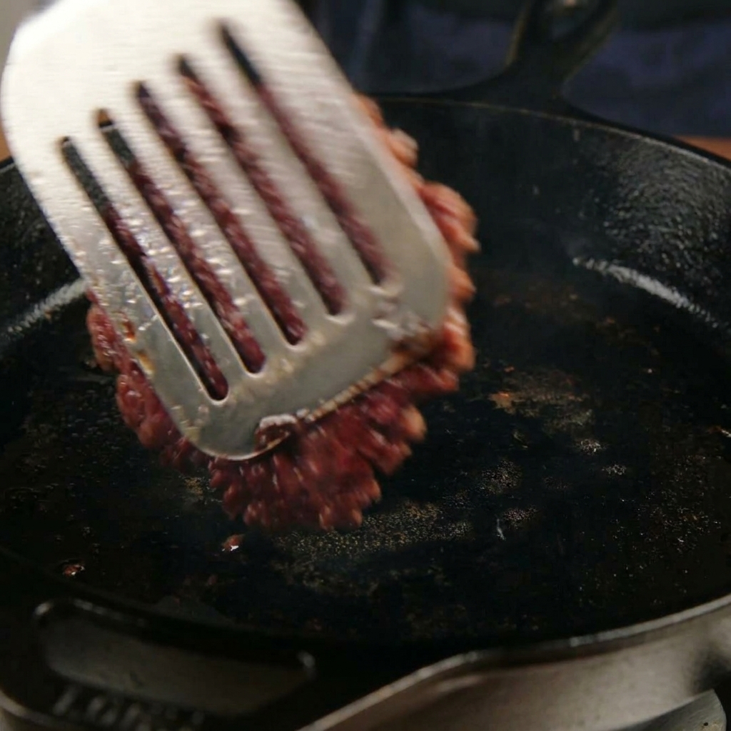 Dando la vuelta a un medallón de carne caramelizado y aplastado en una sartén con una espátula de metal.