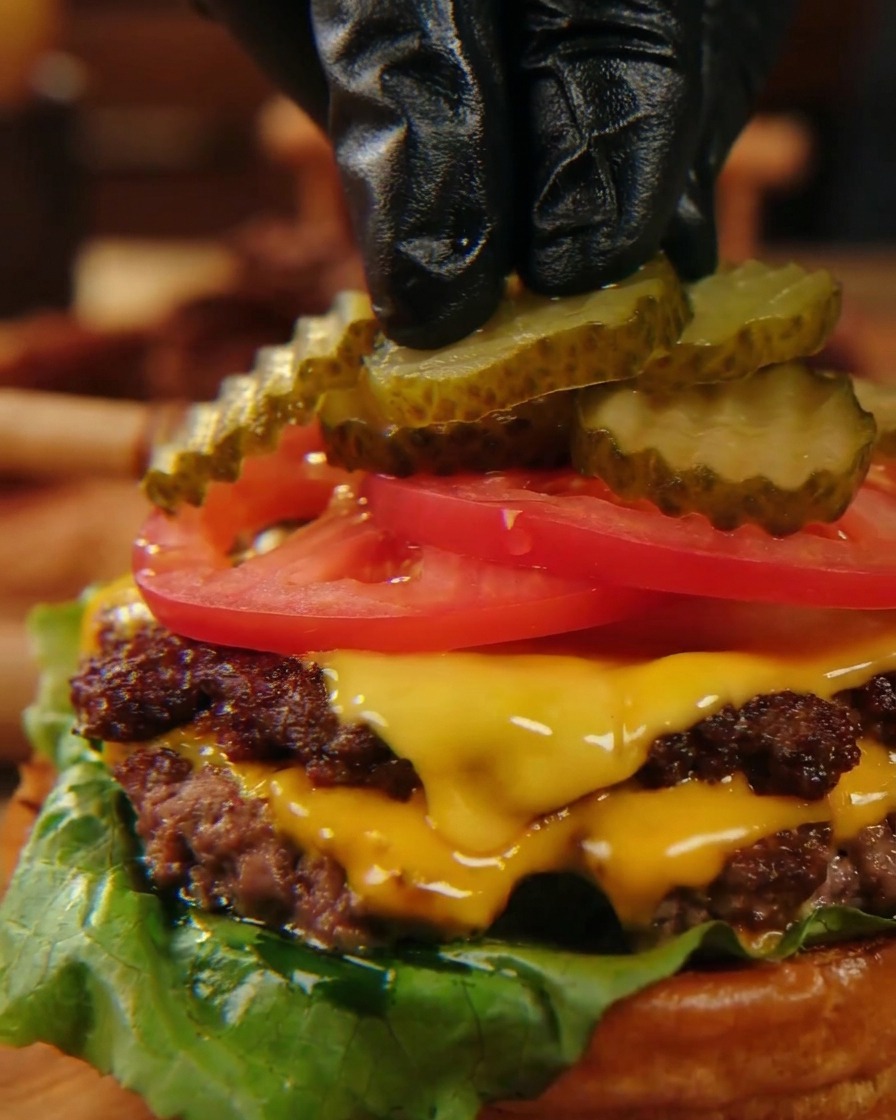 Dedos con guantes negros colocando cuidadosamente rodajas de pepinillo ondulado sobre rodajas de tomate de un rojo intenso, en lo alto de una hamburguesa con queso.