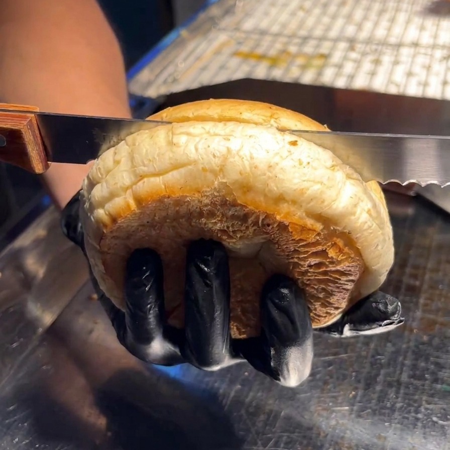 Una persona usando un cuchillo de sierra para cortar un pan de hamburguesa por la mitad sobre una superficie de acero inoxidable.