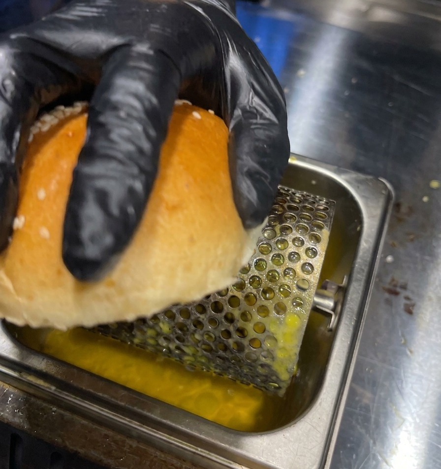 Una mano con guante pasando la cara interna de un pan de hamburguesa por un rodillo de mantequilla en una bandeja de metal.