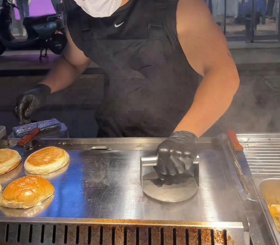 Una prensa metálica circular pesada aplastando una hamburguesa de ternera contra una plancha muy caliente.