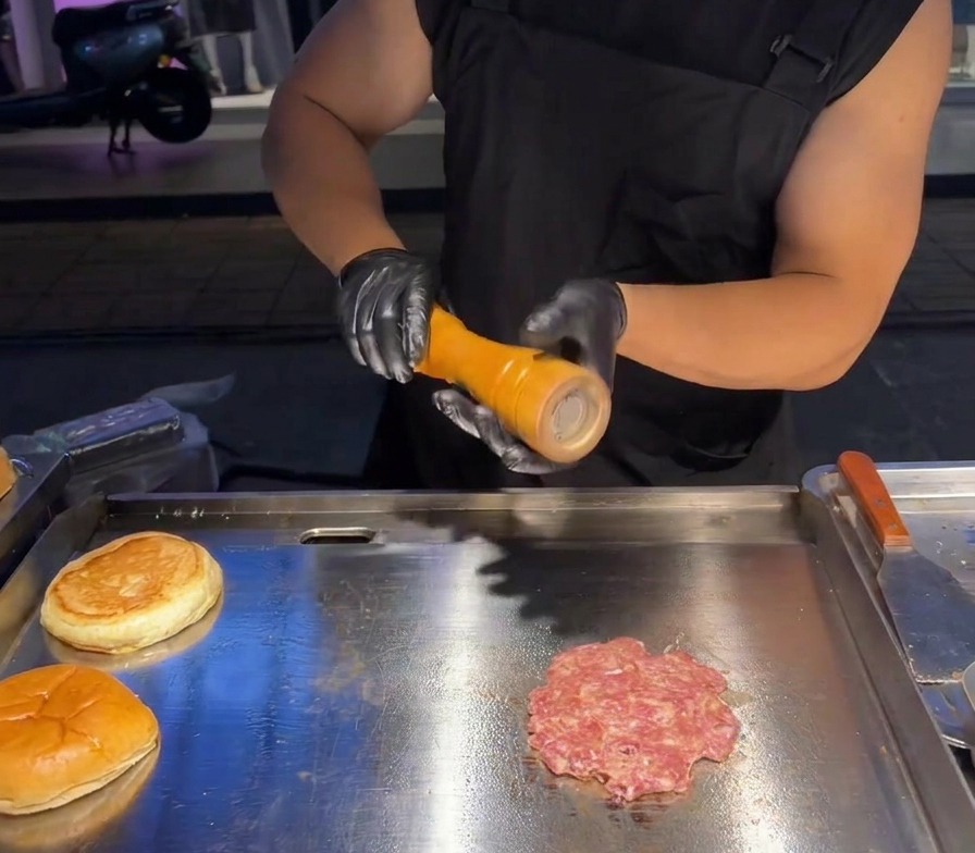 Sal marina recién molida cayendo sobre una hamburguesa aplastada en una plancha profesional.