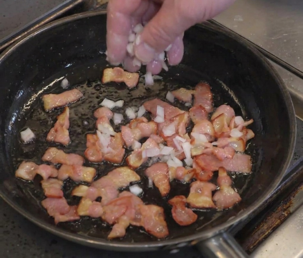 Cebolla picada esparcida sobre trozos de bacón en una sartén.