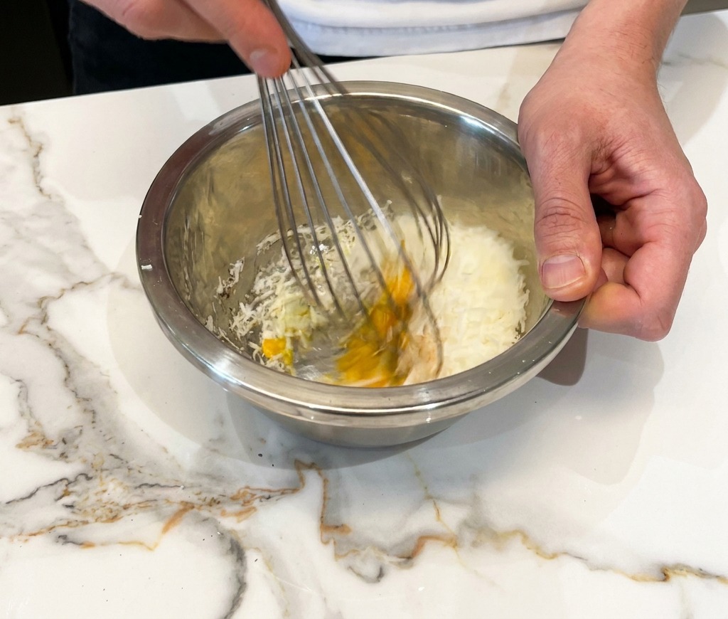 Batiendo huevo, queso rallado y pimienta hasta formar una pasta espesa en un bol metálico.