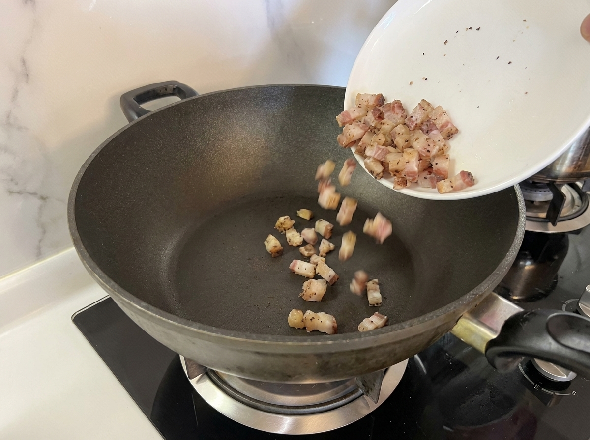 Dados de guanciale crudo vertiéndose desde un plato blanco a una sartén oscura y seca.