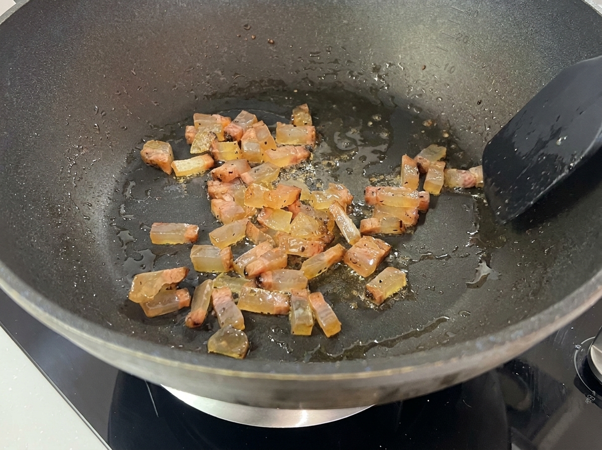 Dados de guanciale friéndose en su propia grasa en una sartén oscura, volviéndose translúcidos y dorados.