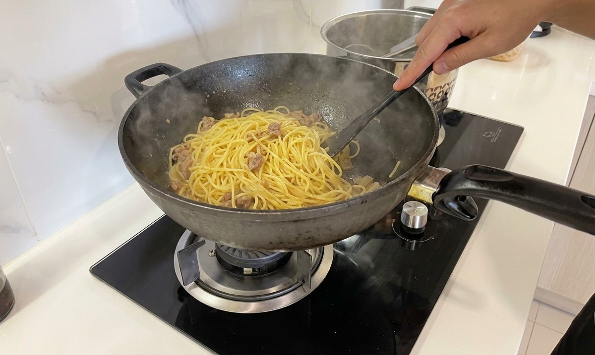 Una mano removiendo y salteando enérgicamente los espaguetis en la sartén para crear una carbonara cremosa.