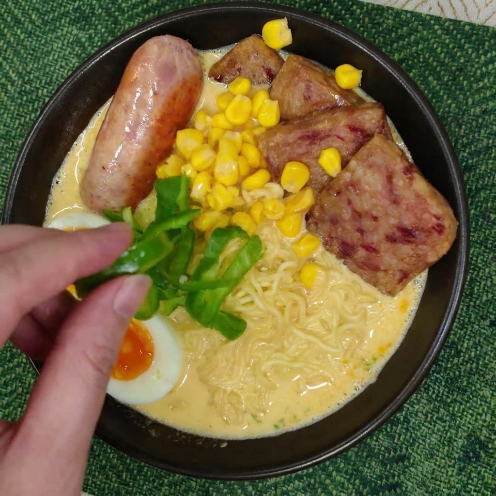 Una mano colocando tiras de pimiento verde fresco sobre un bol de ramen con maíz y carne.