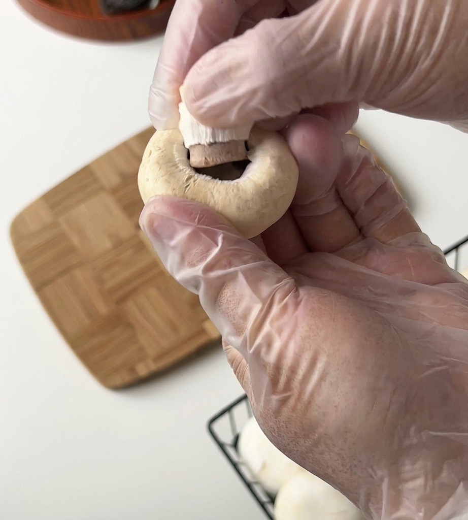 Una persona con guantes retira el tallo de un champiñón blanco sobre una tabla de madera.