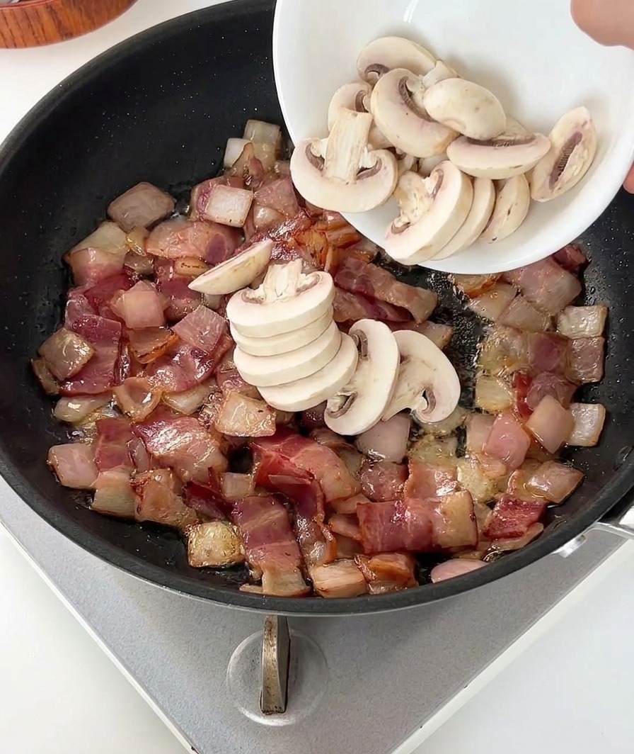 Una persona vertiendo champiñones laminados en la sartén con bacon y cebolla.