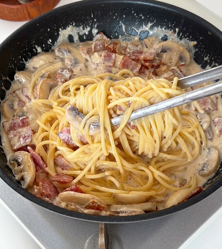 Pinzas mezclando los espaguetis con la salsa espesa, el bacon y los champiñones.