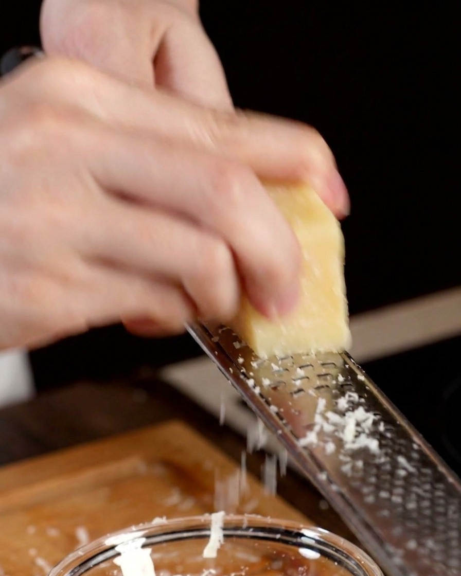 Una mano usando un rallador metálico para desmenuzar un trozo de queso parmesano sobre un bol.