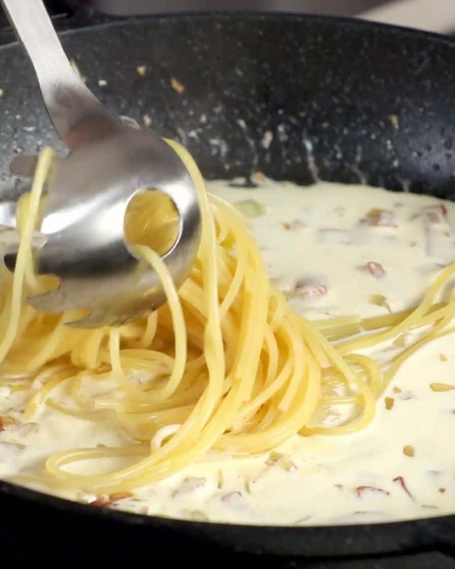 Unas pinzas de pasta pasando espaguetis cocidos directamente a la sartén con la salsa de nata.