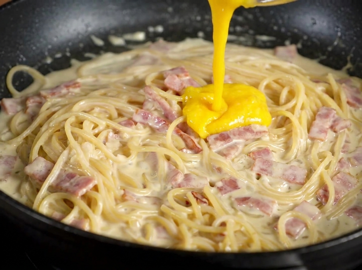 Mezcla espesa de yema de huevo y queso vertiéndose sobre la pasta cremosa en la sartén.