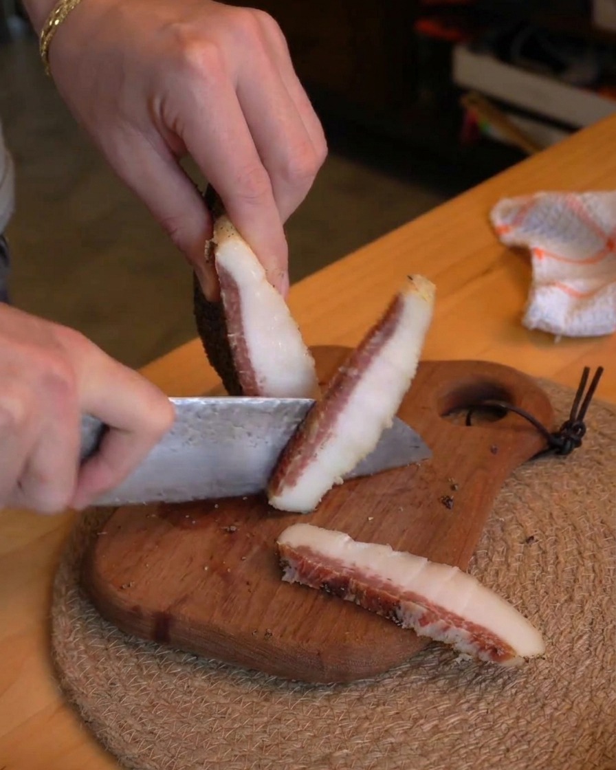 Una persona usando un cuchillo de chef para cortar trozos gruesos de guanciale curado en bastoncillos pequeños sobre una tabla de madera rústica.