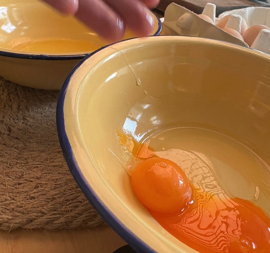 Un primer plano de una yema de huevo de color naranja brillante cayendo en un bol de esmalte amarillo, con cáscaras de huevo visibles al fondo.