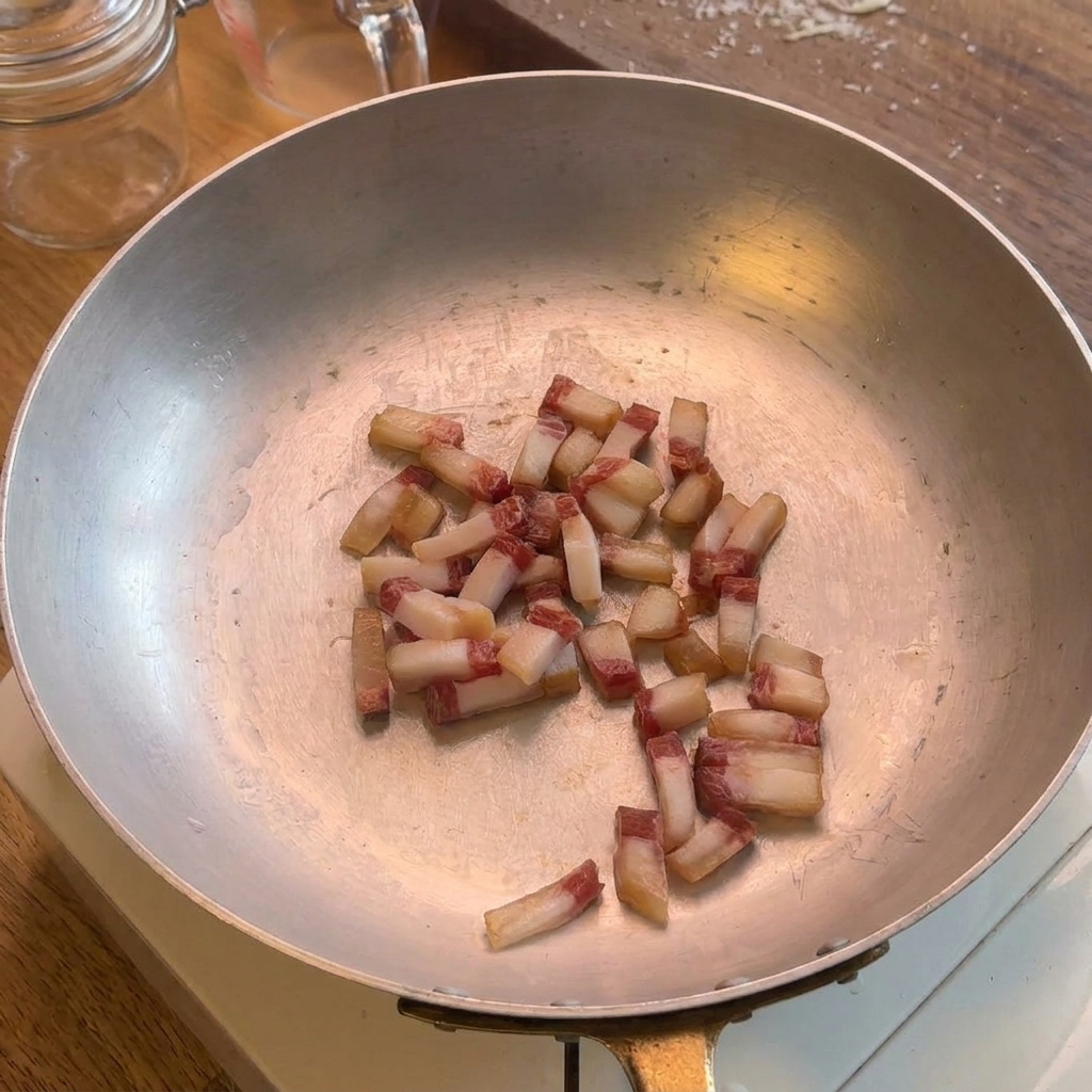 Pequeños cubos de guanciale friéndose en una sartén amplia de acero inoxidable, mostrando la grasa natural empezando a soltarse.