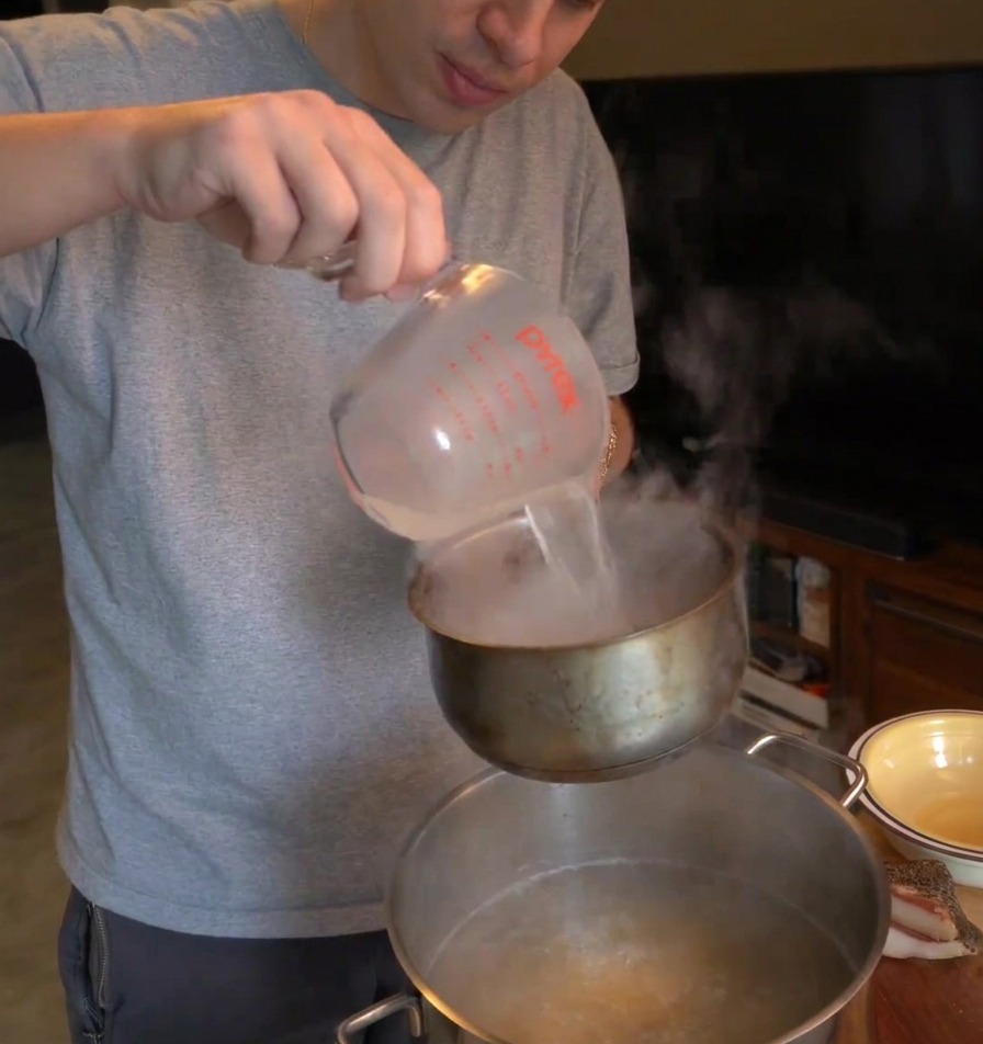Una persona vertiendo agua caliente y turbia de la pasta desde una taza medidora en un cazo pequeño sostenido sobre el agua hirviendo.