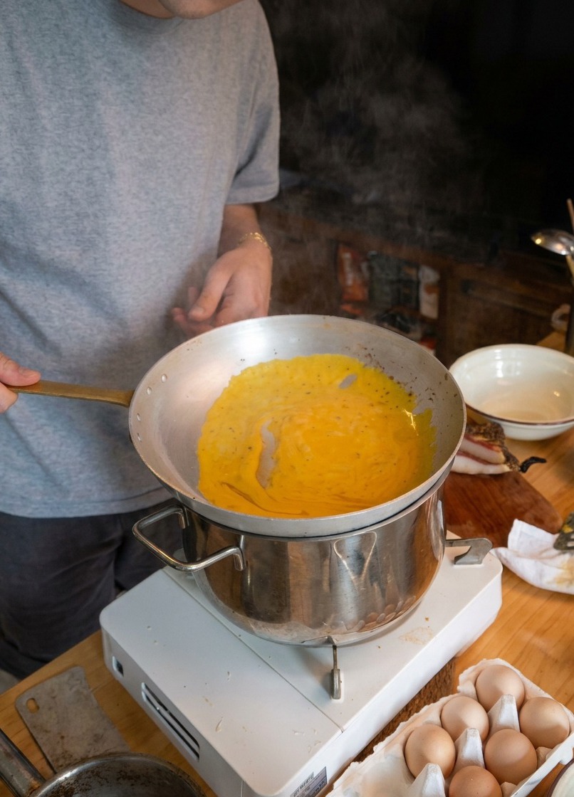 Un chef salteando espaguetis en una sartén de metal ligera sostenida sobre una olla de agua humeante para emulsionar la salsa.