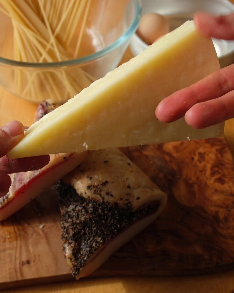 Primer plano de una mano sosteniendo una cuña de queso Pecorino Romano pálido sobre dos trozos gruesos de guanciale con costra de pimienta negra.