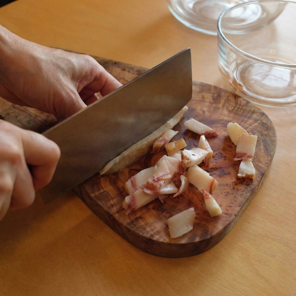 Una persona usando un cuchillo de cocina grande para cortar tiras de guanciale graso sobre una tabla de cortar de madera rústica.