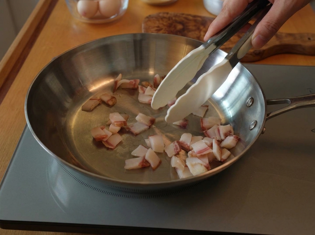 Pinzas removiendo trozos pequeños de guanciale crudo en una sartén ancha de acero inoxidable sobre una placa de inducción.