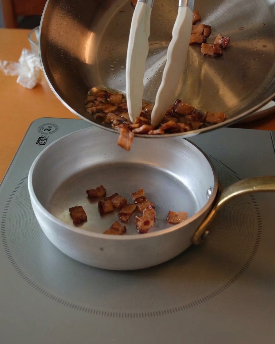 Trozos de guanciale dorados y crujientes vertiéndose desde una sartén de acero inoxidable a un cuenco plateado usando pinzas.