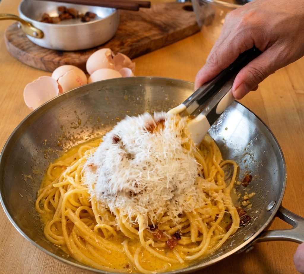 Pinzas removiendo espaguetis en una sartén mientras se añade una gran montaña de queso Pecorino Romano rallado a la crema de yema.