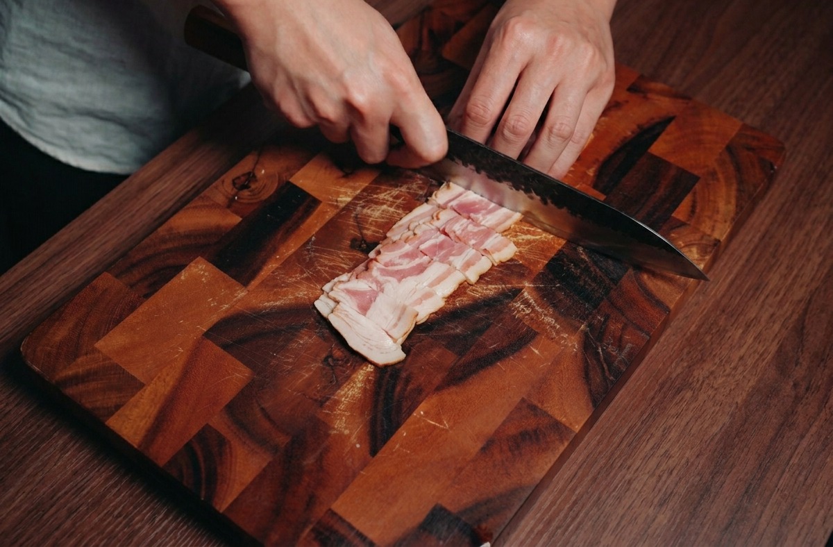 Una persona usando un cuchillo de chef para cortar tiras de beicon en cuadrados pequeños sobre una tabla de madera.