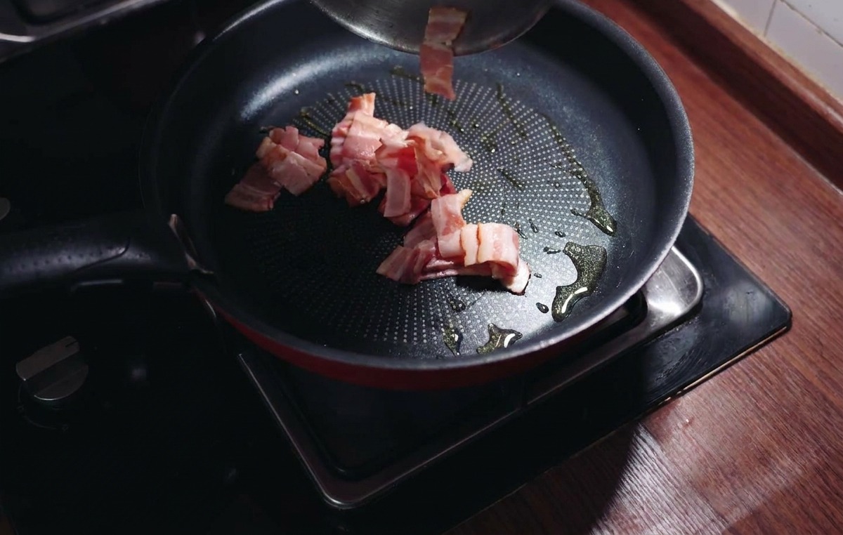 Trozos de beicon picado añadiéndose a una sartén caliente con aceite de oliva brillante.