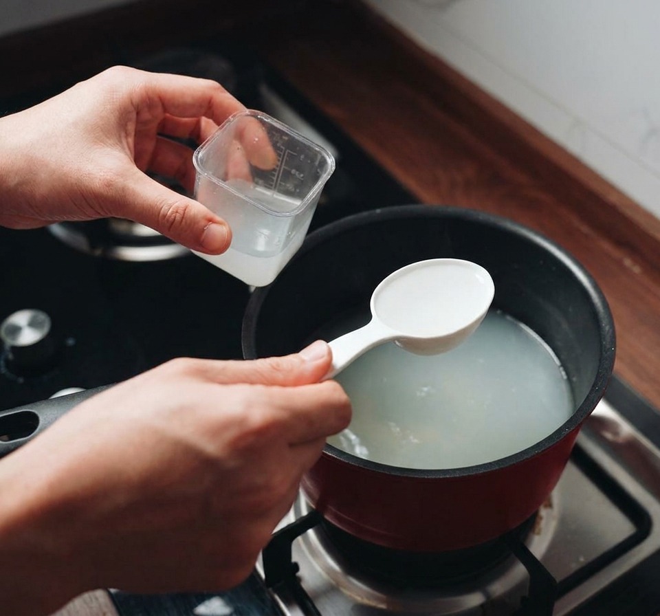 Una persona usando un cazo blanco para sacar agua de pasta turbia de una olla roja en el fuego.