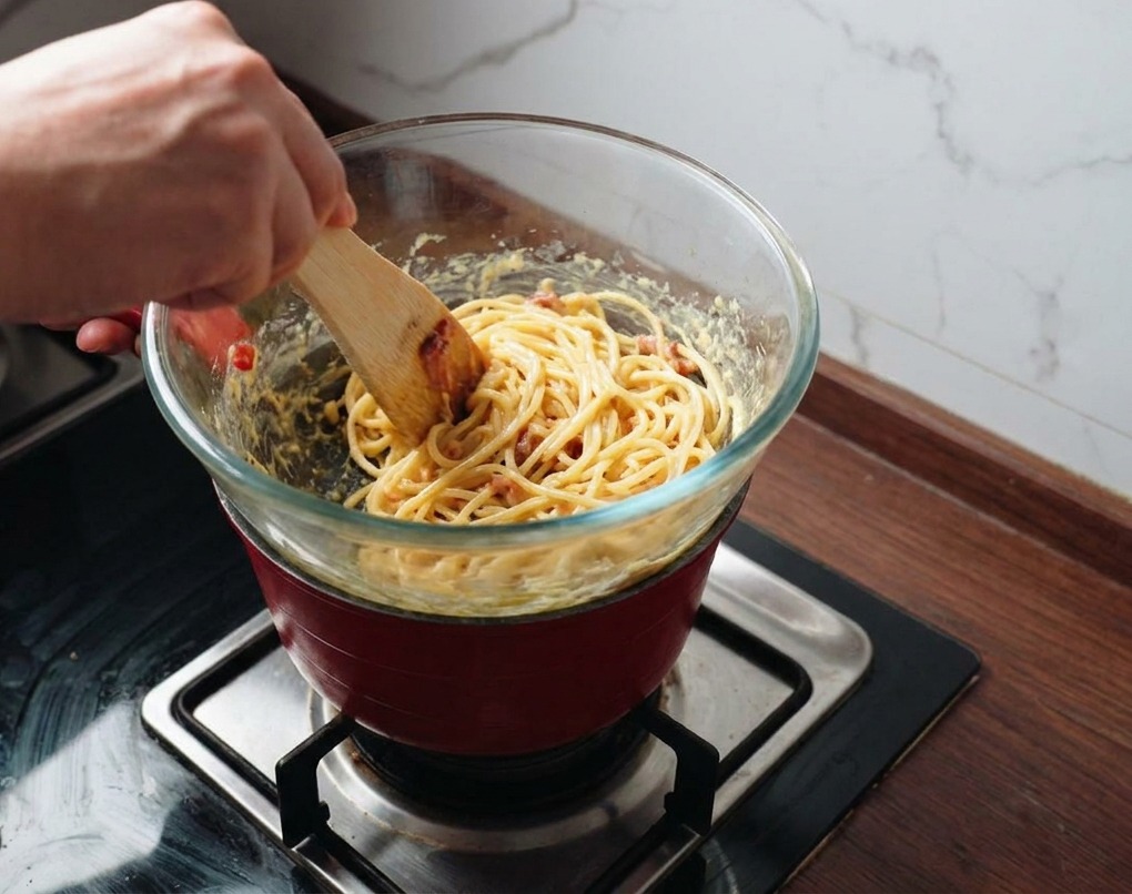Una persona usando una espátula para mezclar pasta y beicon en una salsa dorada sobre una olla humeante.