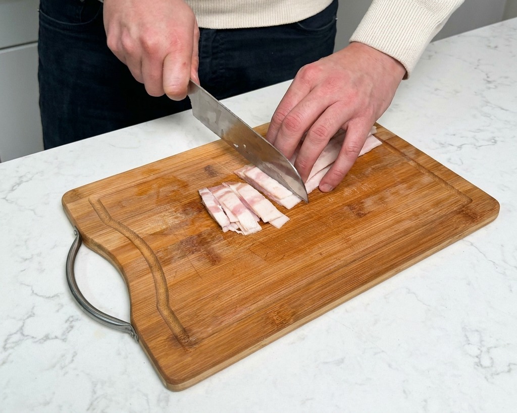 Mano cortando tiras de bacon crudo en pequeños rectángulos sobre una tabla de cortar de madera.