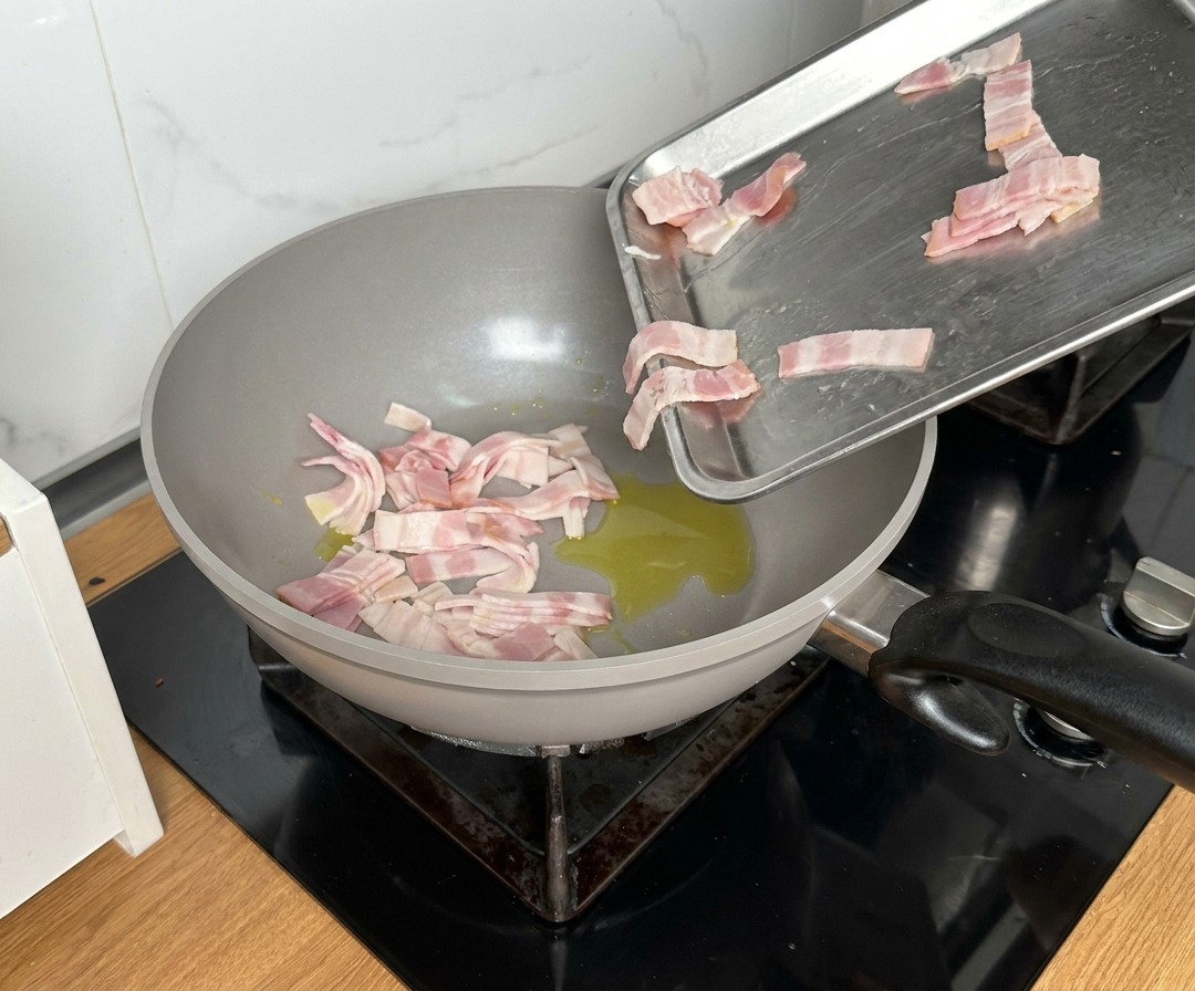 Trozos de bacon cayendo desde una bandeja metálica a una sartén grande con aceite caliente.