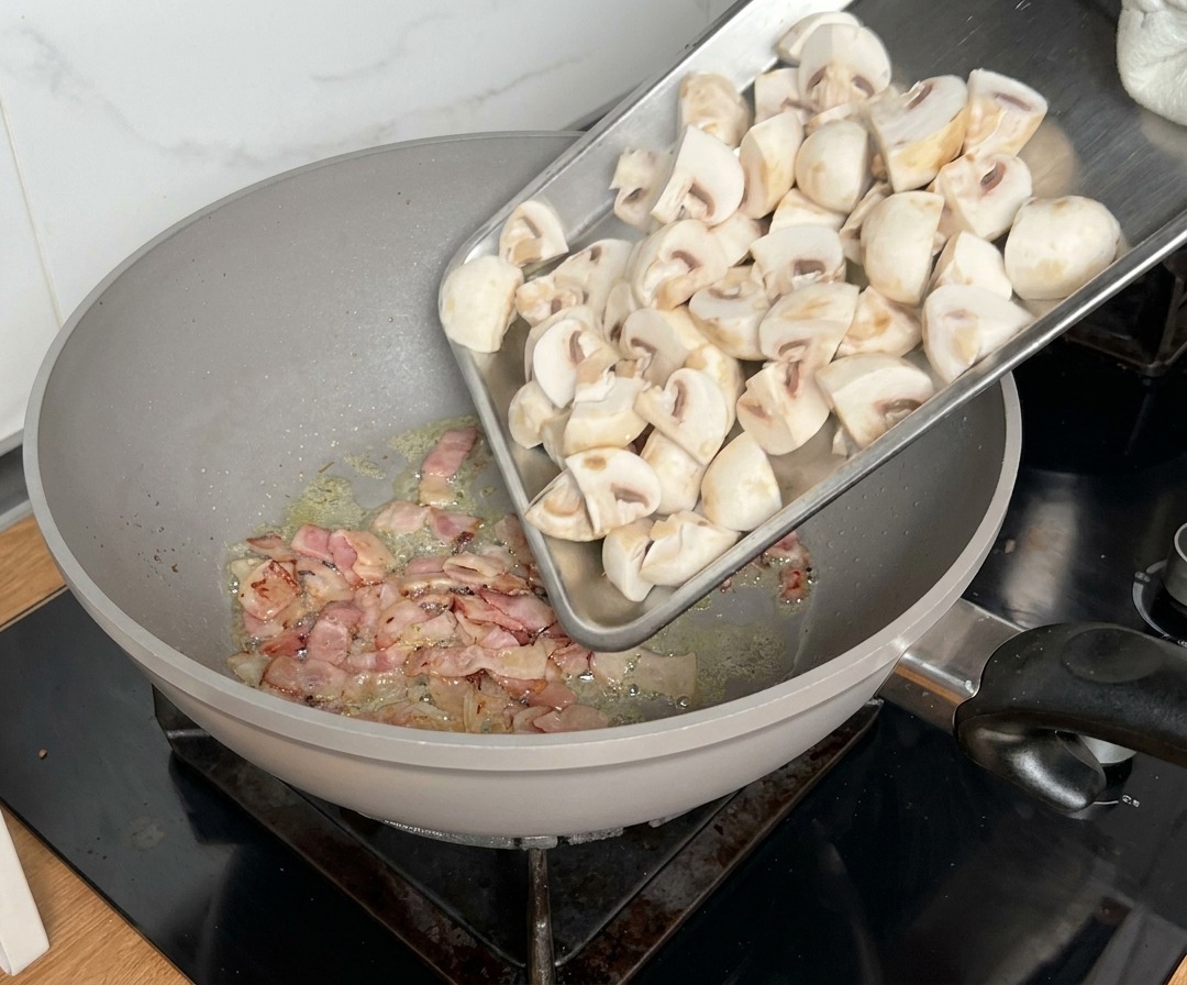 Champiñones cortados en cuartos añadiéndose a una sartén donde el bacon ya se está cocinando.