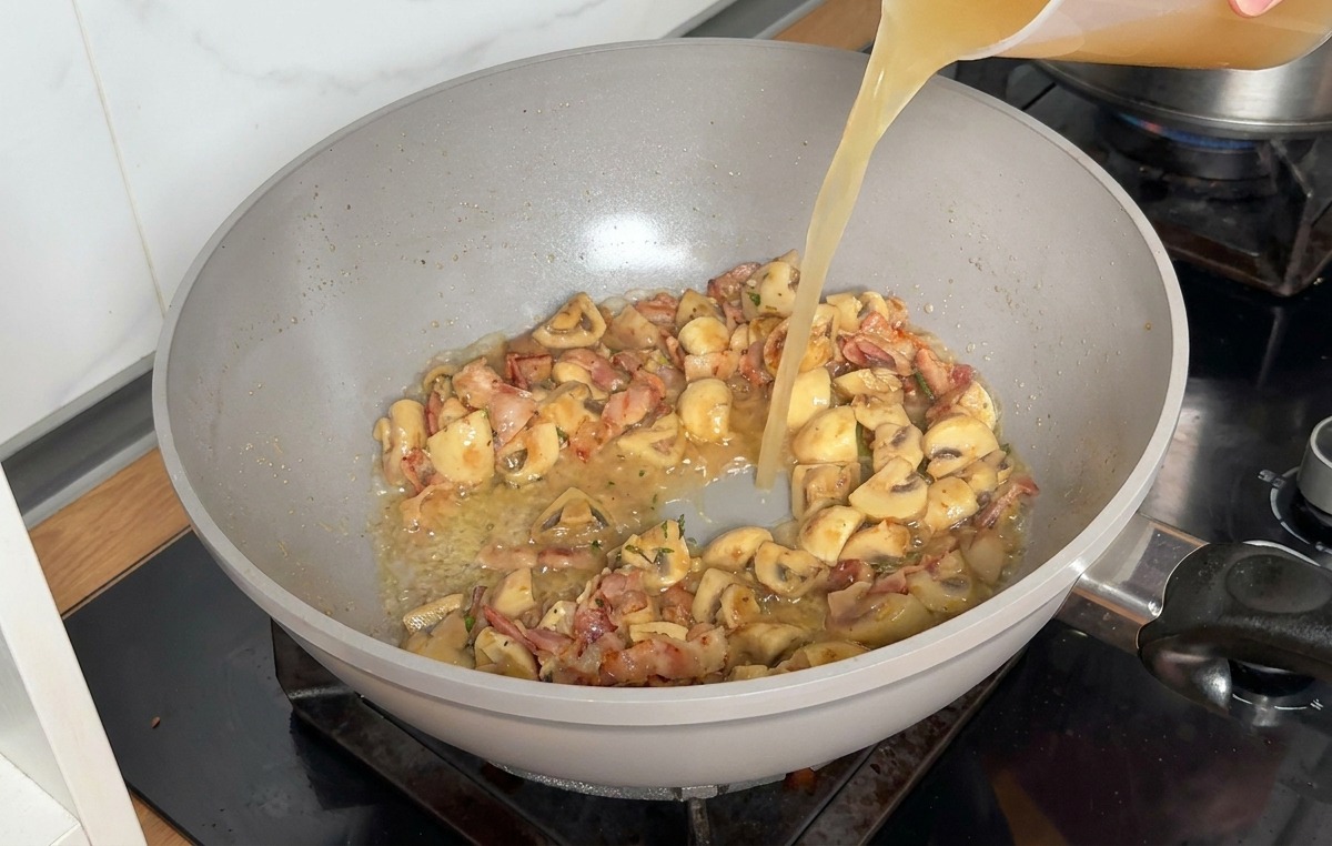 Caldo de pollo vertiéndose desde una jarra medidora de plástico transparente en la sartén con los ingredientes salteados.