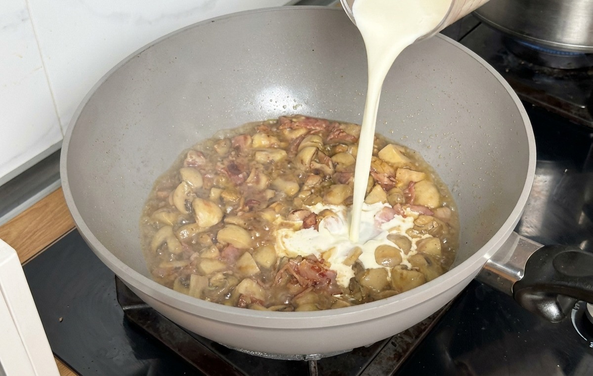 Nata para cocinar vertiéndose desde una jarra medidora en una sartén con caldo reducido y champiñones.