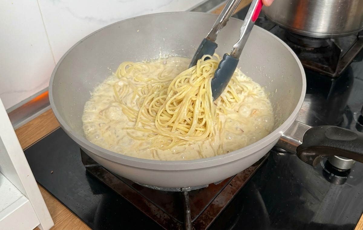 Pasta larga cocida siendo transferida con pinzas metálicas desde una olla a una sartén con salsa cremosa.