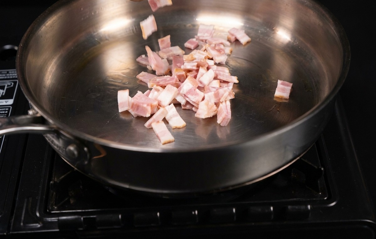 Pequeños trozos rectangulares de bacon cayendo en una sartén metálica brillante.