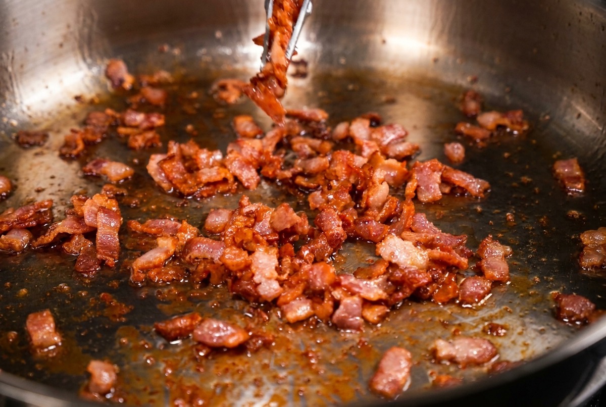 Sartén de acero con trozos de bacon crujientes y marrones siendo retirados con pinzas metálicas.