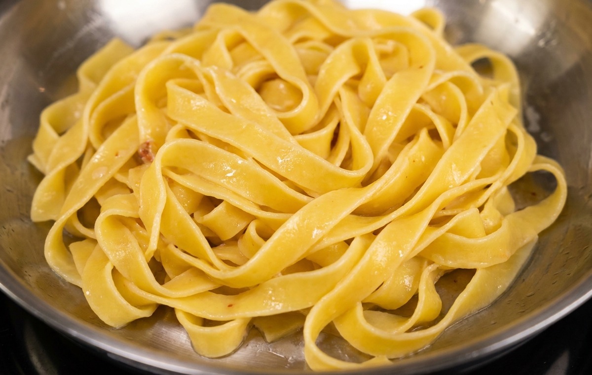 Pasta fettuccine siendo añadida a una sartén de acero con bacon dorado y su grasa.