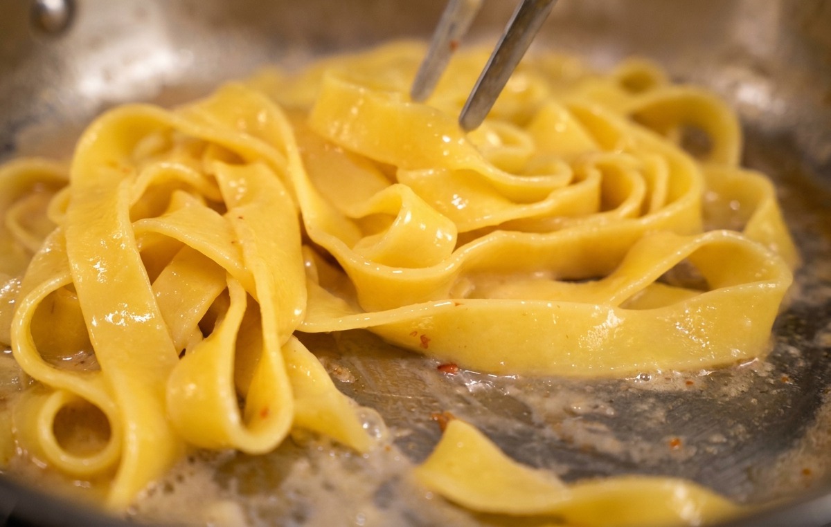 Unas pinzas removiendo pasta larga en una sartén para emulsionar una salsa de huevo amarilla.