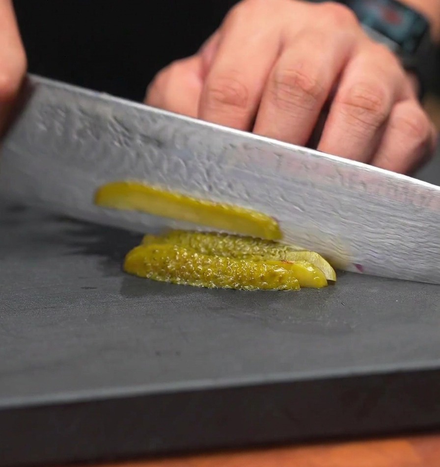 Cuchillo de chef picando pepinillos verdes en trozos muy pequeños y uniformes sobre una superficie de trabajo negra.