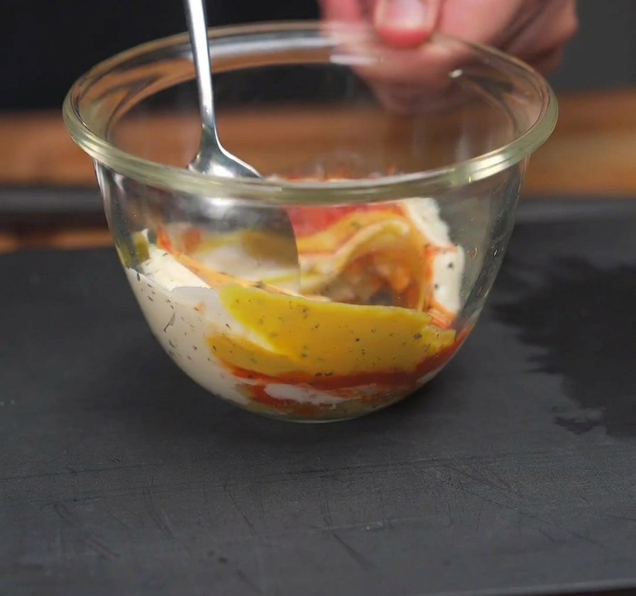 Una cuchara mezclando kétchup rojo, mostaza amarilla y mayonesa blanca en un bol de vidrio transparente.