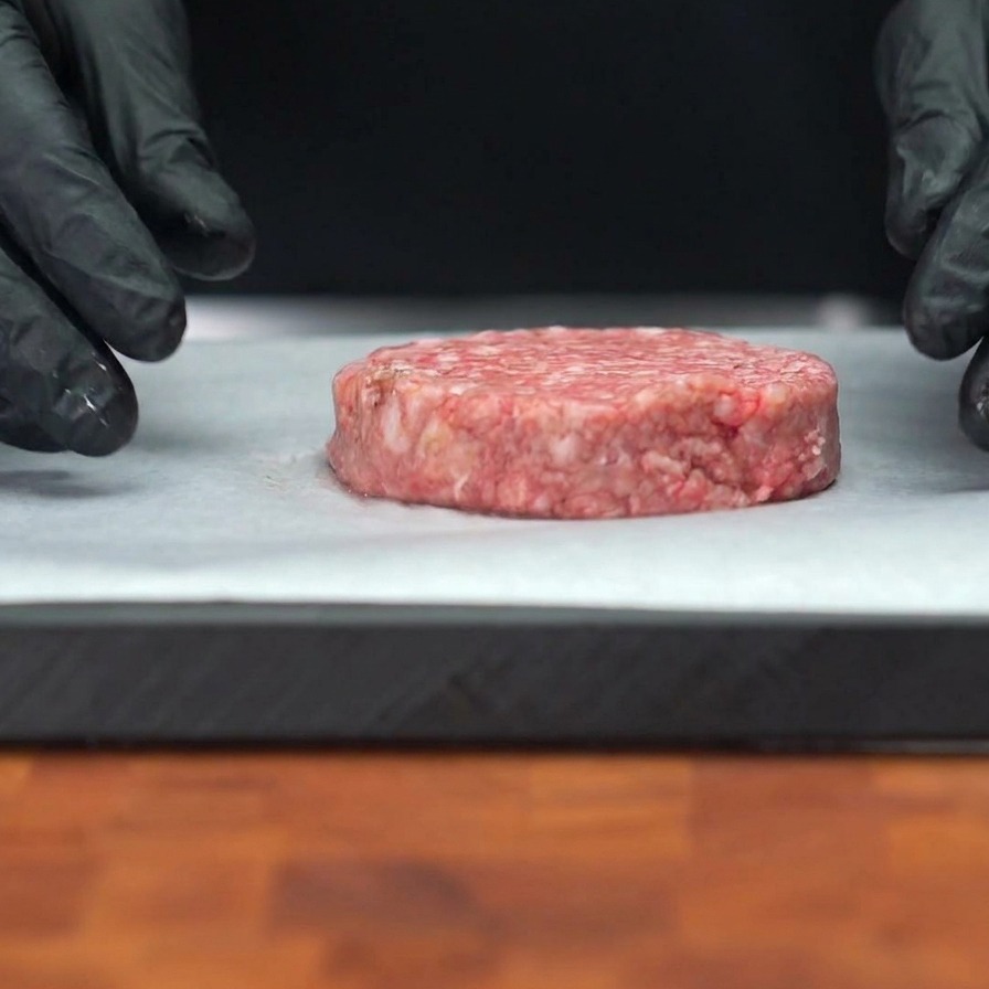 Un medallón grueso de carne de res cruda sobre papel para horno con granos visibles de pimienta negra en la superficie.