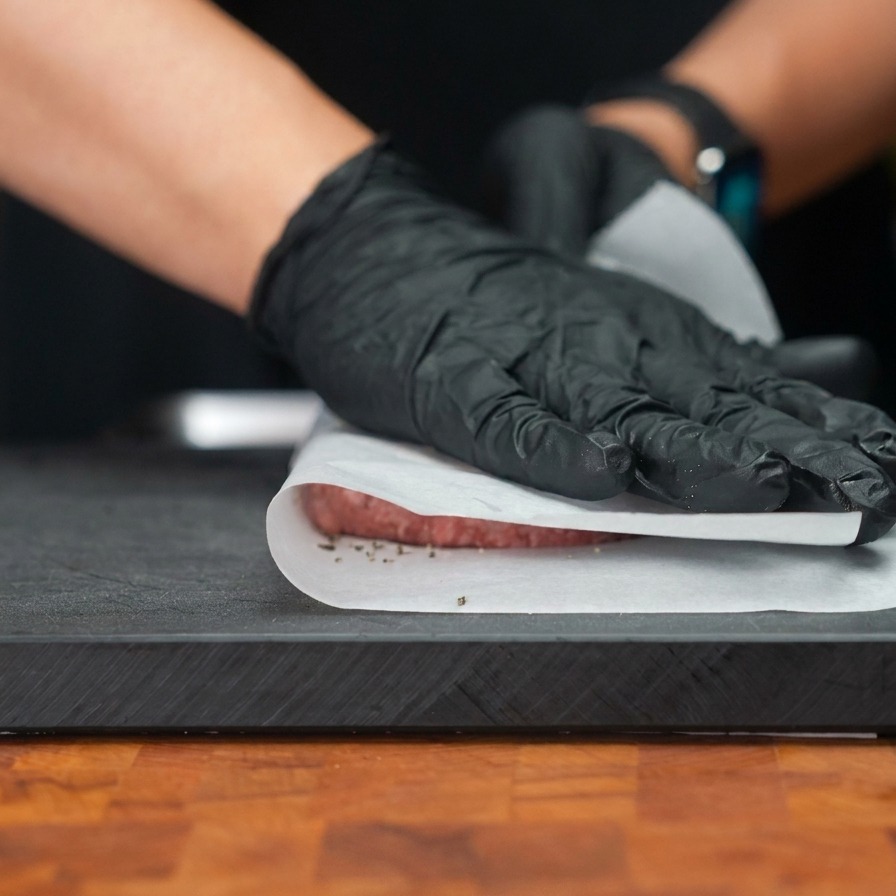 Manos con guantes presionando un medallón de carne a través de una capa de papel para horno para aplanarlo en un disco ancho y fino.
